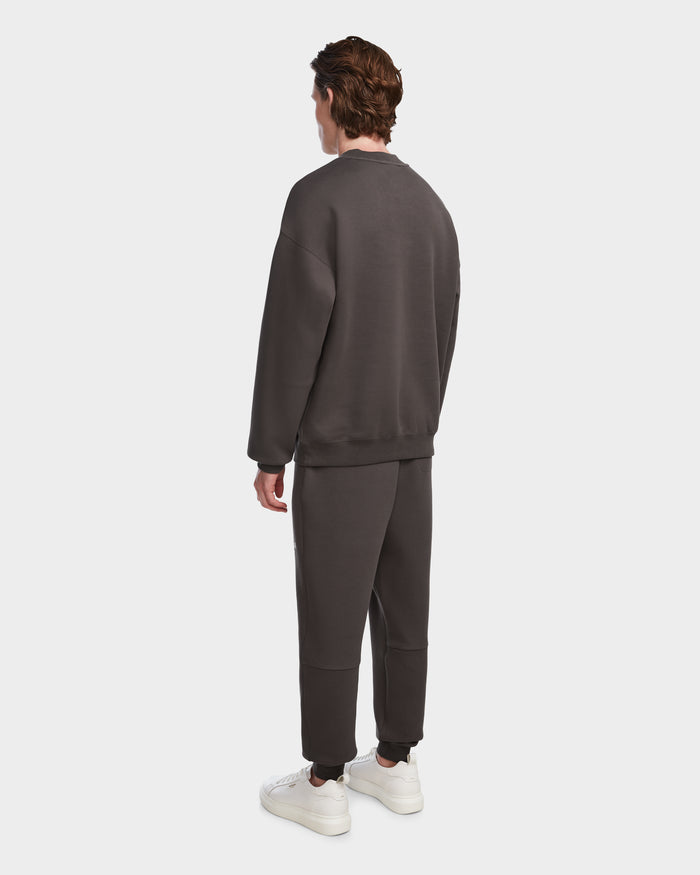 A person with brown hair stands facing away, wearing the Rudsak BRYN - 9524007 Grey M cotton crewneck sweatshirt and matching pants, styled with white sneakers—highlighting timeless design and genderless sizing against a plain white background.