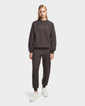A woman with straight blonde hair wears the Rudsak BRYN - 9524007 Grey W cotton crewneck sweatshirt and jogger pants, paired with white sneakers, standing against a plain white background.