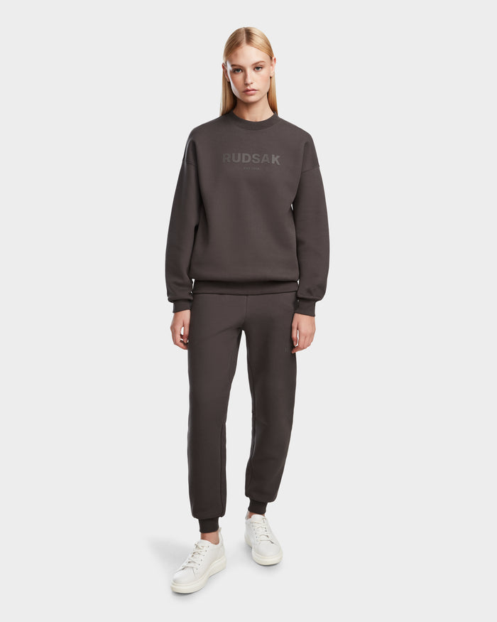 A woman with straight blonde hair wears the Rudsak BRYN - 9524007 Grey W cotton crewneck sweatshirt and jogger pants, paired with white sneakers, standing against a plain white background.