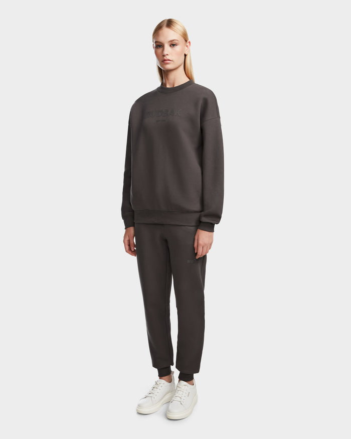 A woman with straight blonde hair wears the Rudsak BRYN - 9524007 Grey W crewneck sweatshirt and matching joggers in genderless sizing, paired with white sneakers, standing against a plain light background.