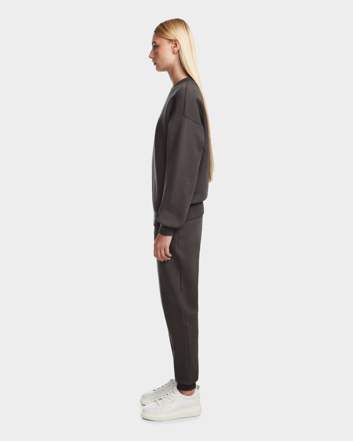 A person with long blond hair stands in profile, wearing the Rudsak BRYN - 9524007 Grey W cotton crewneck sweatshirt and matching pants with white sneakers, showcasing a casual look against a plain white background.