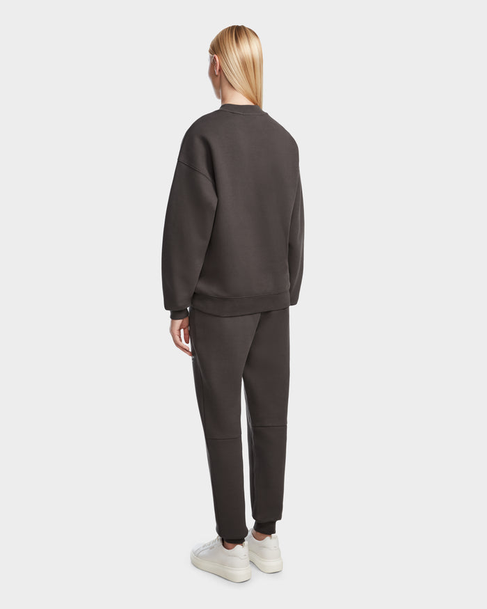 A person with straight, long blond hair faces away, wearing the Rudsak BRYN - 9524007 Grey W crewneck sweatshirt and matching pants in a genderless fit, paired with white sneakers against a plain light background.
