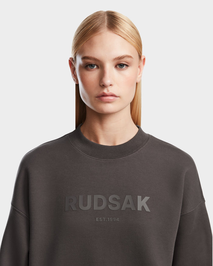 A woman with straight blonde hair wears the Rudsak BRYN - 9524007 Grey W crewneck sweatshirt in dark gray, featuring a cotton build and “RUDSAK EST. 1994” on the front. She stands against a light background, looking at the camera neutrally.