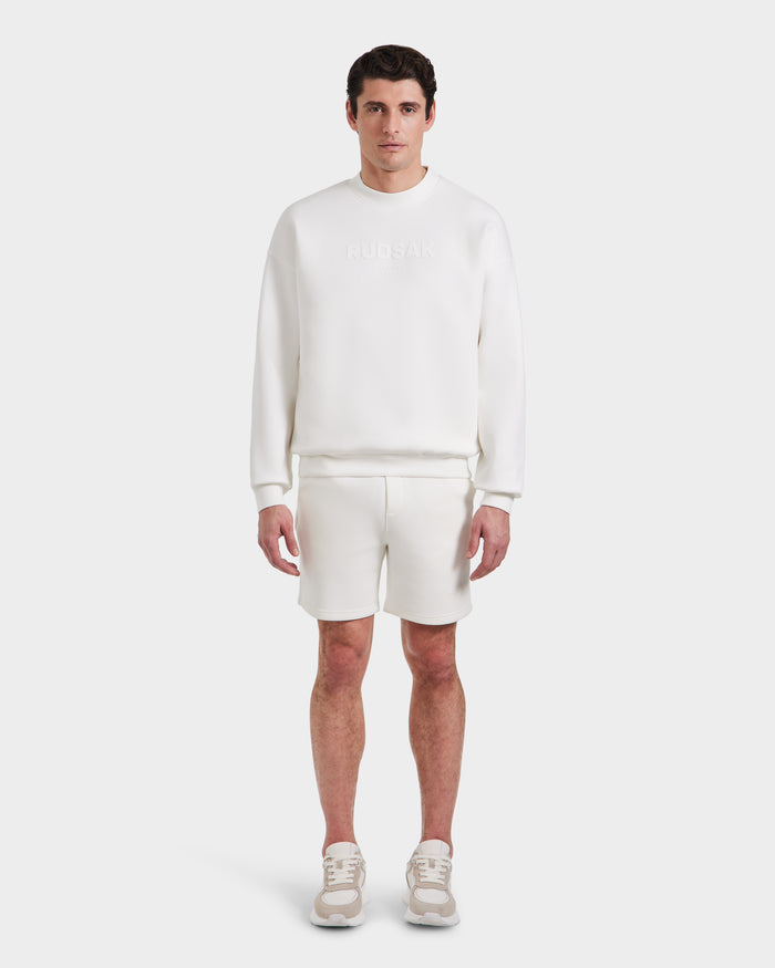 A man faces forward in the Rudsak BRYN - 9524007 Iceland M cotton crewneck sweatshirt with embossed branding, matching shorts, and sneakers. The all-white, genderless outfit delivers an everyday fit against a plain white background.