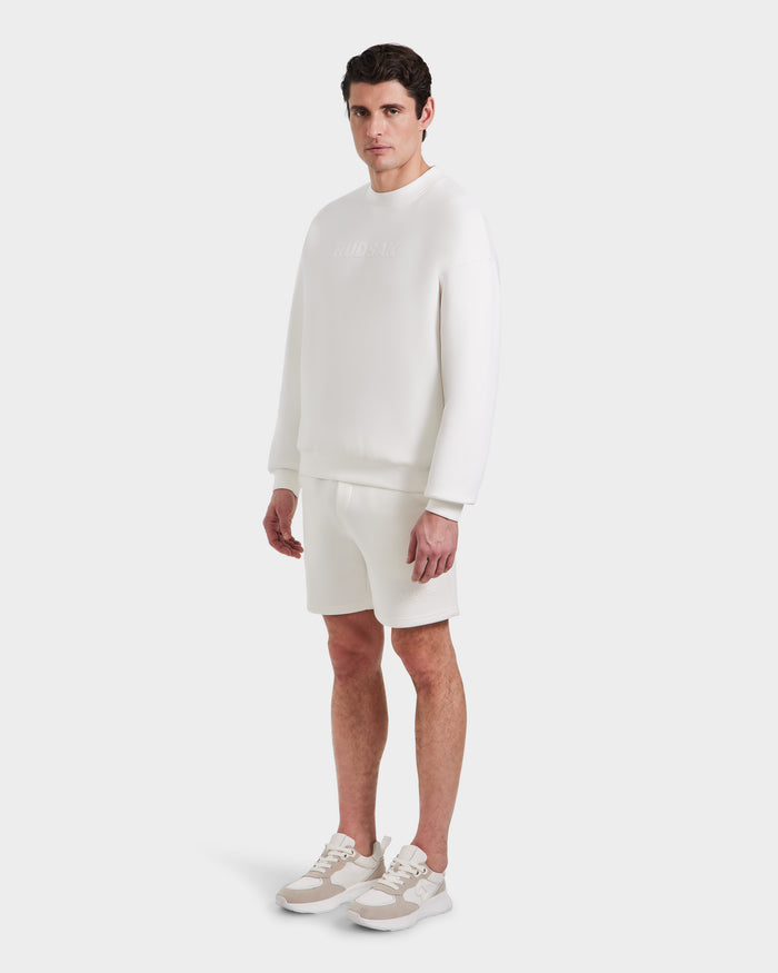A man stands against a plain white background, wearing the Rudsak BRYN - 9524007 Iceland M cotton crewneck sweatshirt, shorts, and sneakers. He faces slightly to the side with a neutral expression.