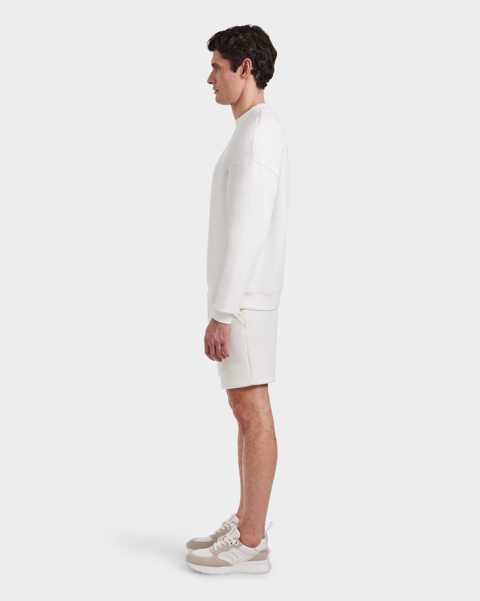 A man in profile faces left, wearing the Rudsak BRYN - 9524007 Iceland M all-white cotton crewneck sweatshirt and shorts set with light beige and white sneakers—an effortless, genderless fit—against a plain white background.