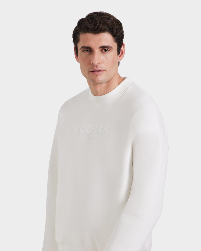 A man with short, dark hair is wearing the Rudsak BRYN - 9524007 Iceland M white crewneck sweatshirt, embossed with the brands logo on the front, standing against a plain white background.