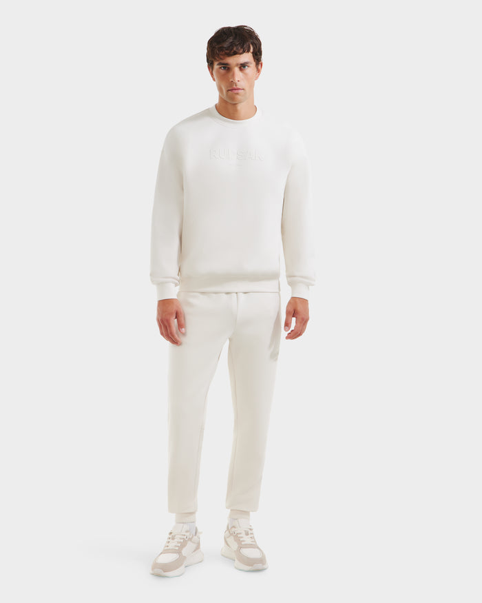 A man faces forward in a casual look, sporting Rudsak’s BRYN - 9524007 Vanilla M crewneck sweatshirt, jogger pants, and beige-and-white sneakers against a plain white background.