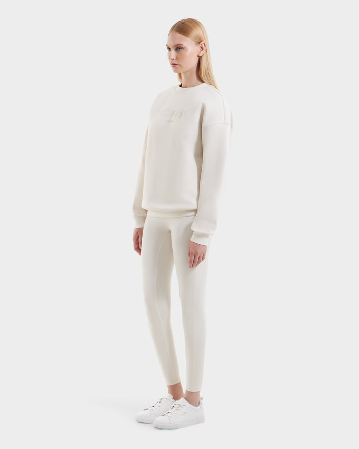 A woman faces slightly to the side, wearing the Rudsak BRYN - 9524007 Vanilla W off-white cotton crewneck sweatshirt with matching leggings and white sneakers, modeling a casual look against a plain white background.