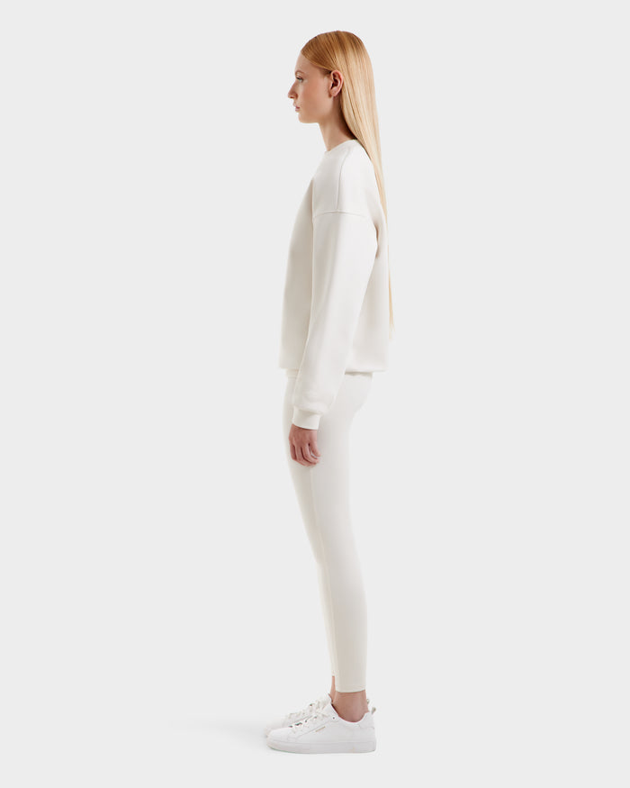 A woman with long straight blonde hair stands in profile, wearing the Rudsak BRYN - 9524007 Vanilla W crewneck sweatshirt in genderless sizing, paired with white leggings and sneakers against a plain white background.