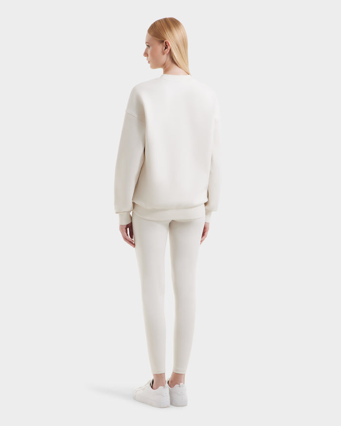 A woman with straight blonde hair faces away, wearing the Rudsak BRYN - 9524007 Vanilla W off-white cotton crewneck sweatshirt, matching leggings, and white sneakers for a comfy everyday fit against a plain light background.