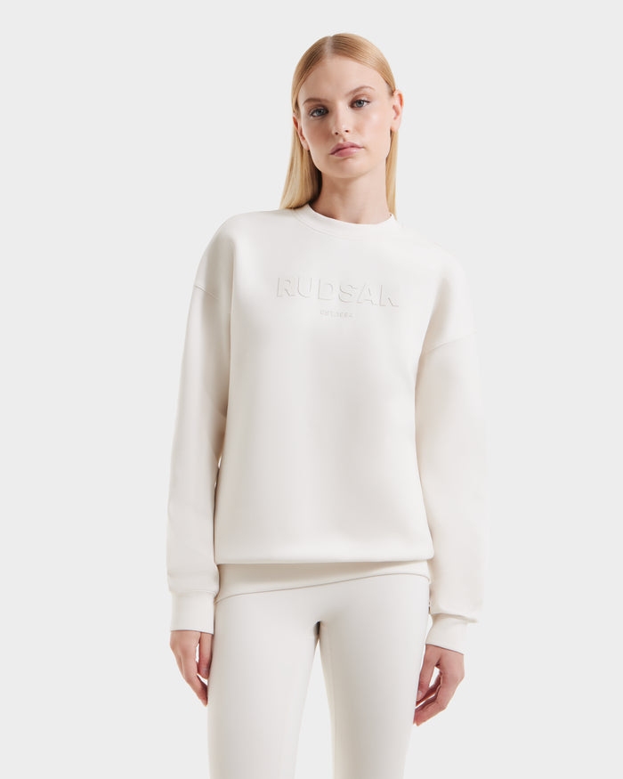 A woman with straight blonde hair wears the Rudsak BRYN - 9524007 Vanilla W off-white cotton crewneck sweatshirt with embossed "RUDSAK" on the front and matching leggings, standing against a plain white background—ideal for everyday wear.