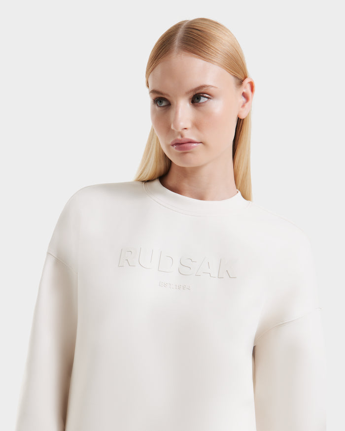 A woman with straight blonde hair wears the Rudsak BRYN - 9524007 Vanilla W cotton crewneck sweatshirt, featuring embossed RUDSAK and EST. 1994 on the front, standing against a plain white background.