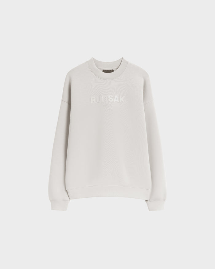 The BRYN - 9524007 Vanilla W by Rudsak is a plain white cotton crewneck sweatshirt with long sleeves, ribbed cuffs, and RUDSAK embroidered across the chest. It features a genderless fit and is shown against a plain white background.