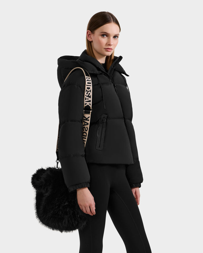 A woman with straight brown hair tucked behind her ears faces left, wearing a black puffer jacket and leggings. She carries the Rudsak CABAS - 8325545 Black faux fur mini tote with a beige strap featuring black text.