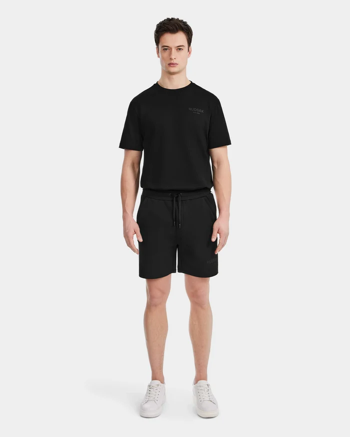 A man faces forward wearing Rudsak’s CAMDEN - 6525028 Black t-shirt and jogger shorts with white sneakers against a plain white background—an ideal look for any casual wardrobe.