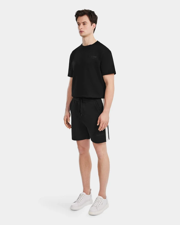 A man stands against a white background wearing Rudsak’s CAMDEN - 6525028 Black cotton-blend men’s jogger shorts, paired with a black t-shirt and white sneakers, showcasing a relaxed and casual wardrobe style.
