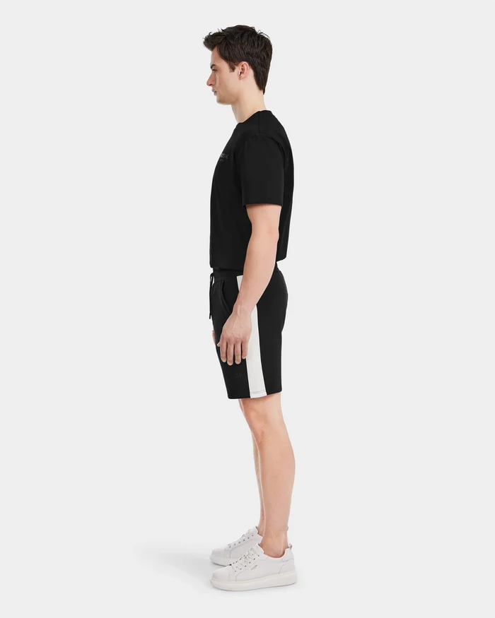 A man stands in profile facing left, wearing Rudsak’s CAMDEN - 6525028 Black cotton-blend men’s jogger shorts with a white side stripe, a black t-shirt, and white sneakers against a plain white background—an effortless casual look.
