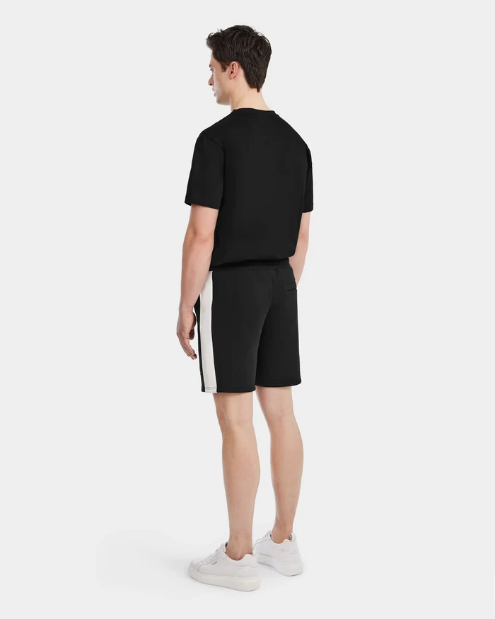 A man stands facing away in the Rudsak CAMDEN - 6525028 Black T-shirt, men’s jogger shorts with white side stripes, and white sneakers against a light background—a laid-back addition to any casual wardrobe.