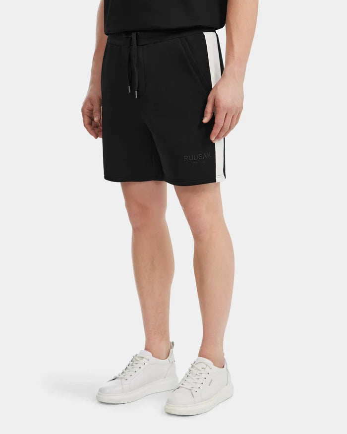 A person wears Rudsak’s CAMDEN - 6525028 black men’s jogger shorts with white side stripes, a black shirt, and white sneakers. The cotton-blend shorts feature “RUDSAK” on the left leg for a casual, relaxed look.