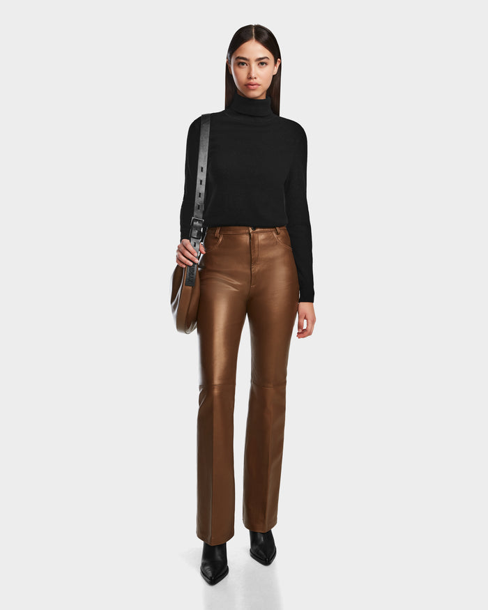 A woman poses against a white background, wearing the Rudsak CARISSA - 8524545 Antique Bronze high-fashion leather pants, paired with a black turtleneck sweater, black pointed shoes, and a black shoulder bag.
