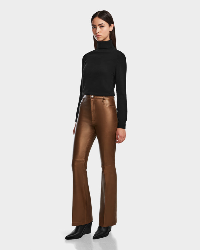 A woman with straight dark hair and a neutral expression models the Rudsak CARISSA - 8524545 Antique Bronze high-fashion flare leather pants, paired with a black turtleneck sweater and black ankle boots against a plain background.