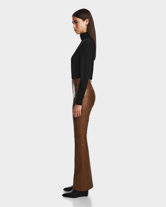 A woman stands in profile against a plain background, wearing the CARISSA - 8524545 Antique Bronze high-waisted flare leather pants by Rudsak, paired with a black long-sleeve turtleneck top and black shoes.