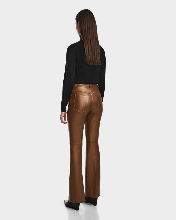 A woman with straight brown hair, wearing a black long-sleeve top and Rudsak CARISSA - 8524545 Antique Bronze leather flare pants, stands facing away on a plain white background.