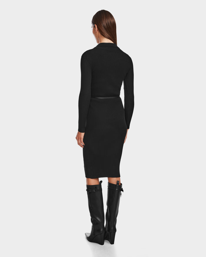 A woman faces away from the camera, wearing the Rudsak CARLI - 8524573 Black fitted knit dress with a vegan leather belt and knee-high black boots, all set against a plain light background.