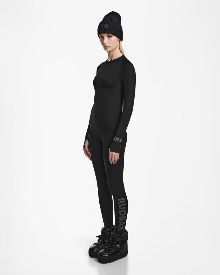 A woman wears the Rudsak CARRIE - 8524619 Black beanie, fitted ski base layer top, stretch leggings with RUDSAK text on the leg, and black winter boots, standing against a white background and looking at the camera neutrally.