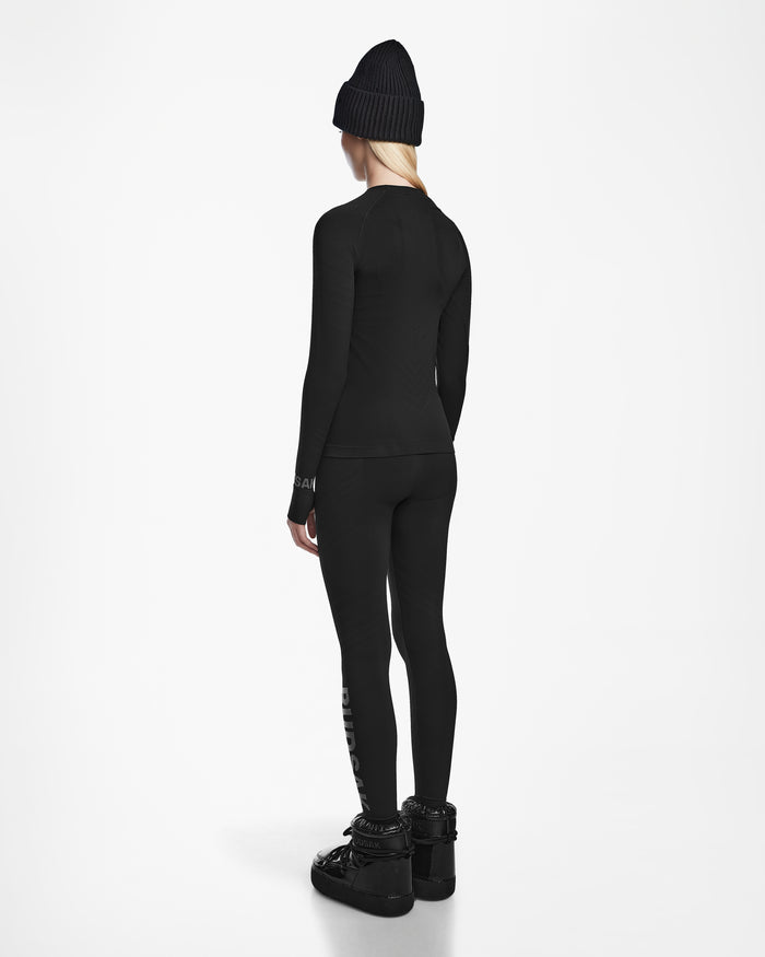 A person with long blonde hair wears the Rudsak CARRIE - 8524619 Black women’s thermal set and black boots, styled with a black beanie, standing back to the camera against a plain white background.