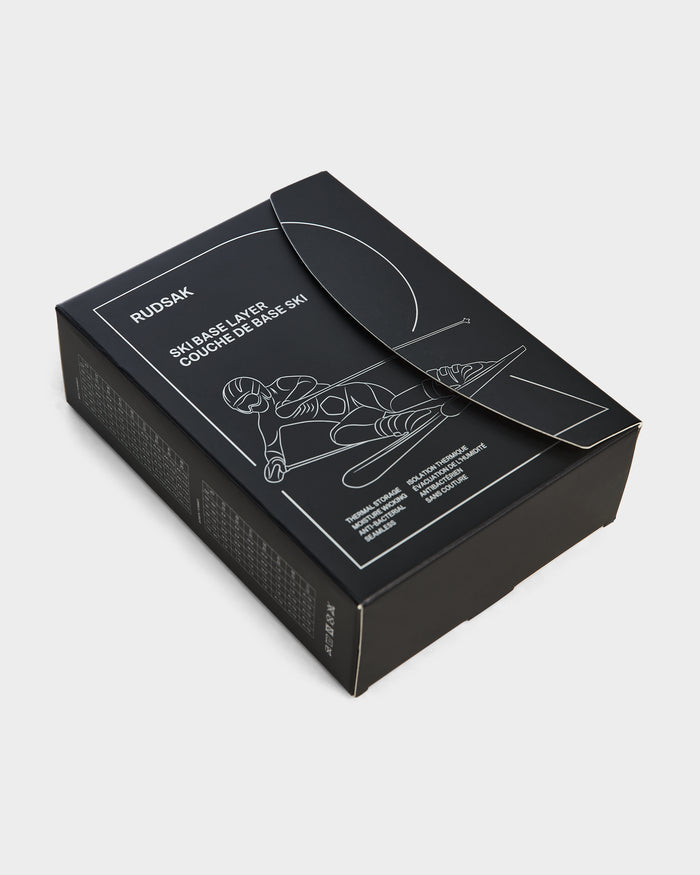 A black rectangular RUDSAK box labeled CARRIE - 8524619 Black features a white skier line drawing and SKIABLE LAYER COUCHE DE BASE SKI text, hinting at a womens thermal set inside. The partially open box is shown on a plain white background.