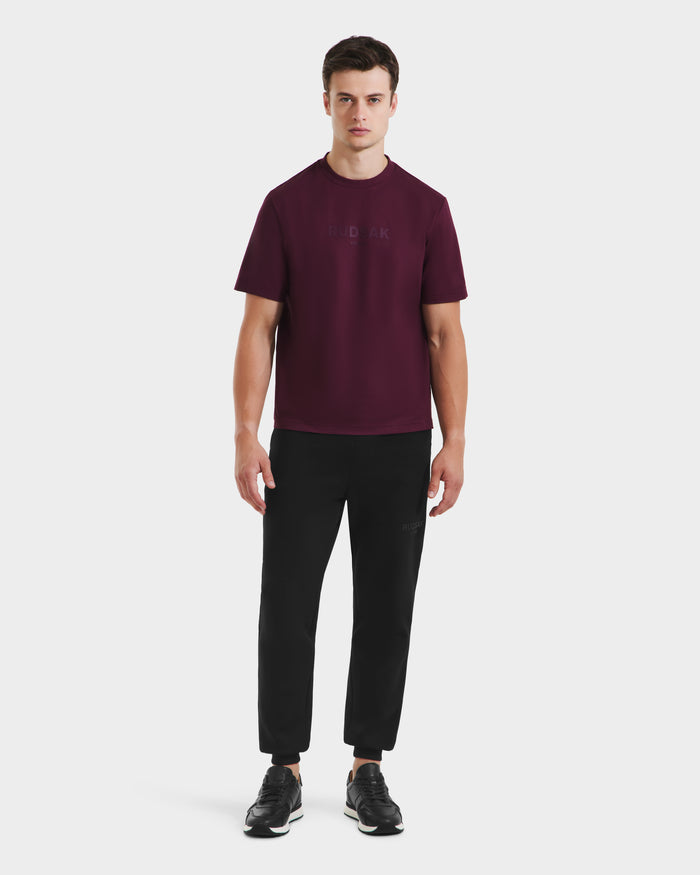 A man faces forward against a plain white background, wearing the Rudsak CASEY - 6525037 Bordeaux T-shirt paired with black pants and black sneakers.