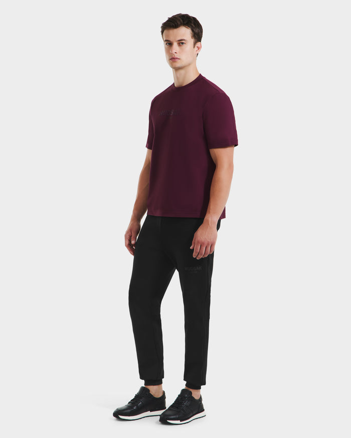 A man stands against a plain white background wearing the Rudsak CASEY - 6525037 Bordeaux T-shirt, black jogger pants, and black sneakers, highlighting this comfortable essential with a relaxed posture and neutral expression.