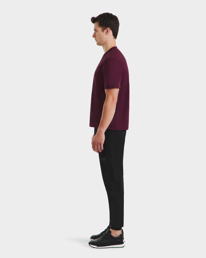 A man stands in profile facing left, wearing a CASEY - 6525037 Bordeaux T-shirt by Rudsak, paired with black pants and black athletic shoes—an effortlessly comfortable essential against a plain white background.