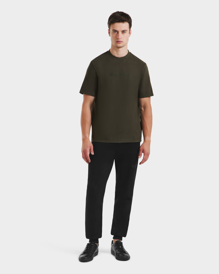 A man faces forward against a plain white background, wearing the Rudsak CASEY - 6525037 dark olive T-shirt with black pants and black sneakers.
