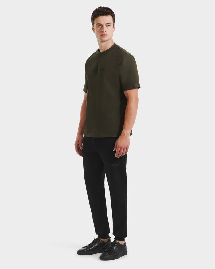 A young man stands slightly angled, wearing a Rudsak CASEY - 6525037 Dark Olive mens T-shirt, black jogger pants, and black sneakers against a plain white background.