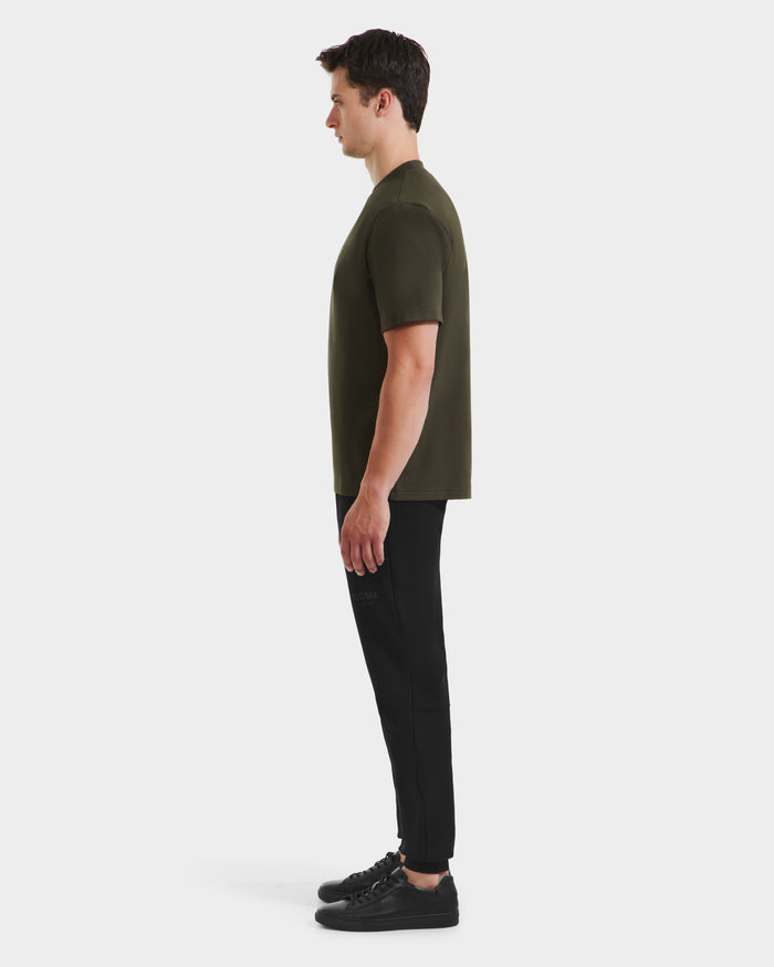 A man stands in profile facing left, wearing a Rudsak CASEY - 6525037 T-shirt in dark olive, with black pants and black sneakers, set against a plain white background.
