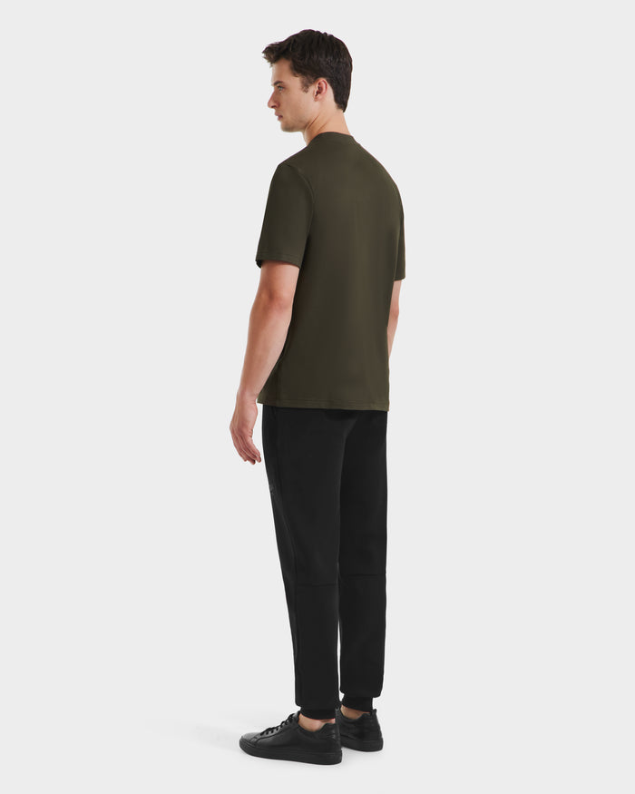 A man faces away, wearing the Rudsak CASEY - 6525037 Dark Olive mens T-shirt, black pants, and black sneakers against a plain white background.