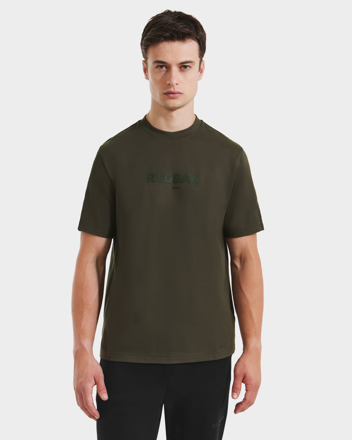 A young man faces forward in the Rudsak CASEY - 6525037 Dark Olive men's T-shirt with chest print, paired with black pants against a plain white background.
