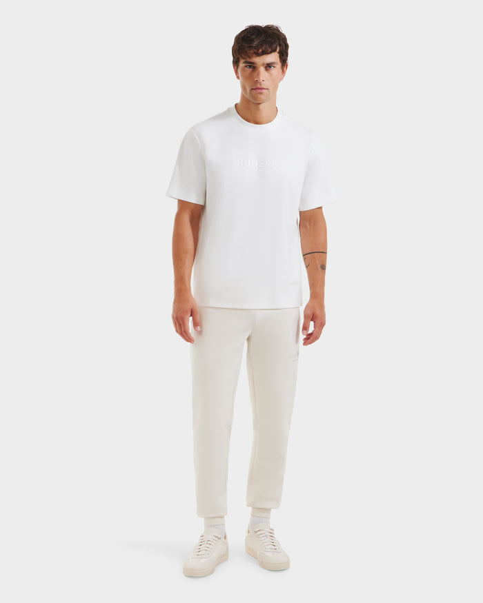 A man with short brown hair and a neutral expression faces forward in the Rudsak CASEY - 6525037 Optic White T-shirt, styled with white pants and sneakers, set against a plain white background.