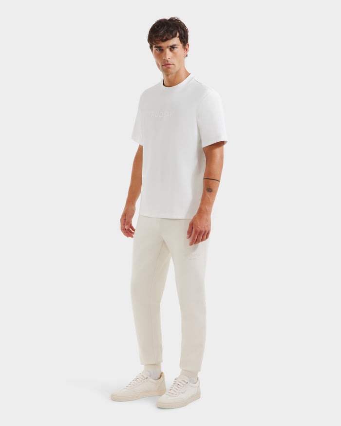 A man with short dark hair and a neutral expression stands against a plain white background, wearing the Rudsak CASEY - 6525037 Optic White T-shirt, light beige jogger pants, and white sneakers, with one arm relaxed by his side.