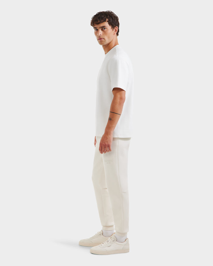 A man stands sideways in a classic Rudsak CASEY - 6525037 Optic White men’s T-shirt, paired with white jogger pants and white sneakers against a plain light background.