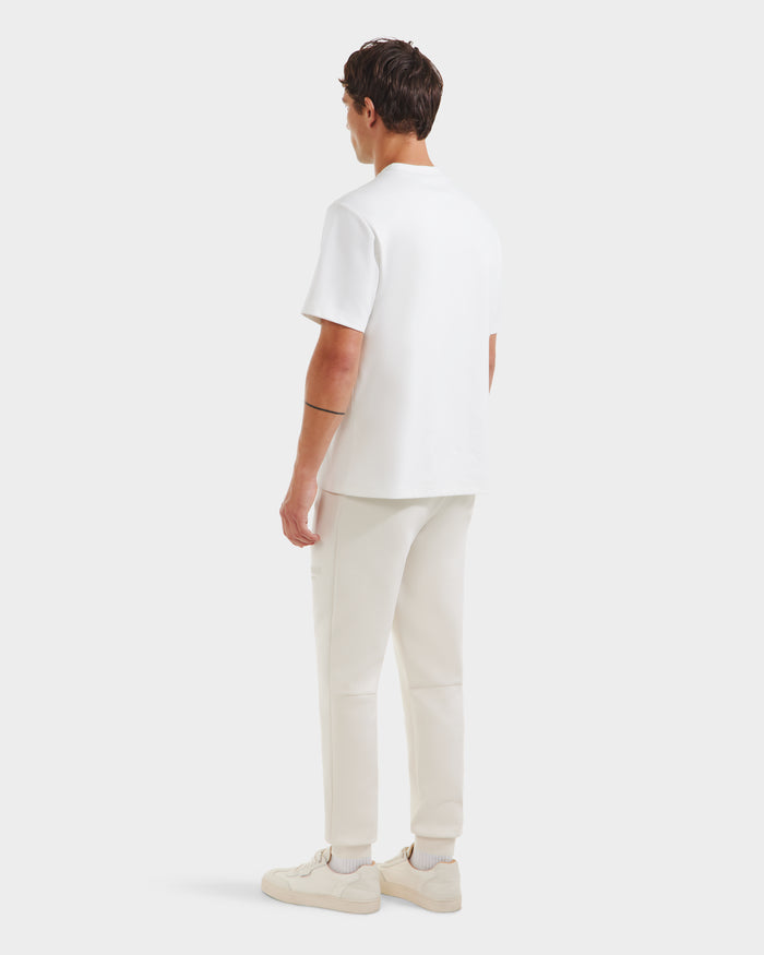 A person faces away from the camera, wearing a Rudsak CASEY - 6525037 Optic White T-shirt, white pants, and white sneakers against a light gray background.
