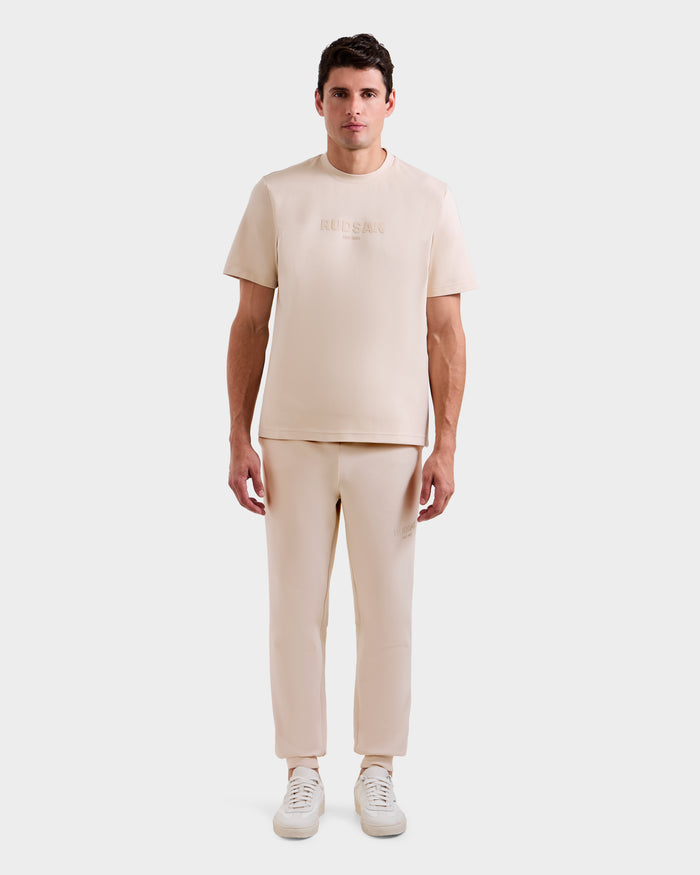 A man faces forward in a Rudsak CASEY - 6525037 Vanilla men’s T-shirt, paired with beige pants and white sneakers, against a plain white background.