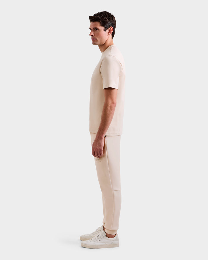A man stands in profile against a plain background, wearing the Rudsak CASEY - 6525037 Vanilla mens T-shirt and pants set, paired with white sneakers.