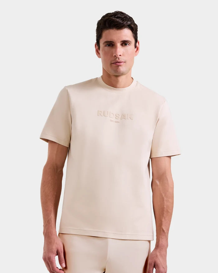 A man with short dark hair wears the Rudsak CASEY - 6525037 Vanilla men’s T-shirt with matching pants, standing against a plain light grey background. The T-shirt has subtle embroidered text across the chest.