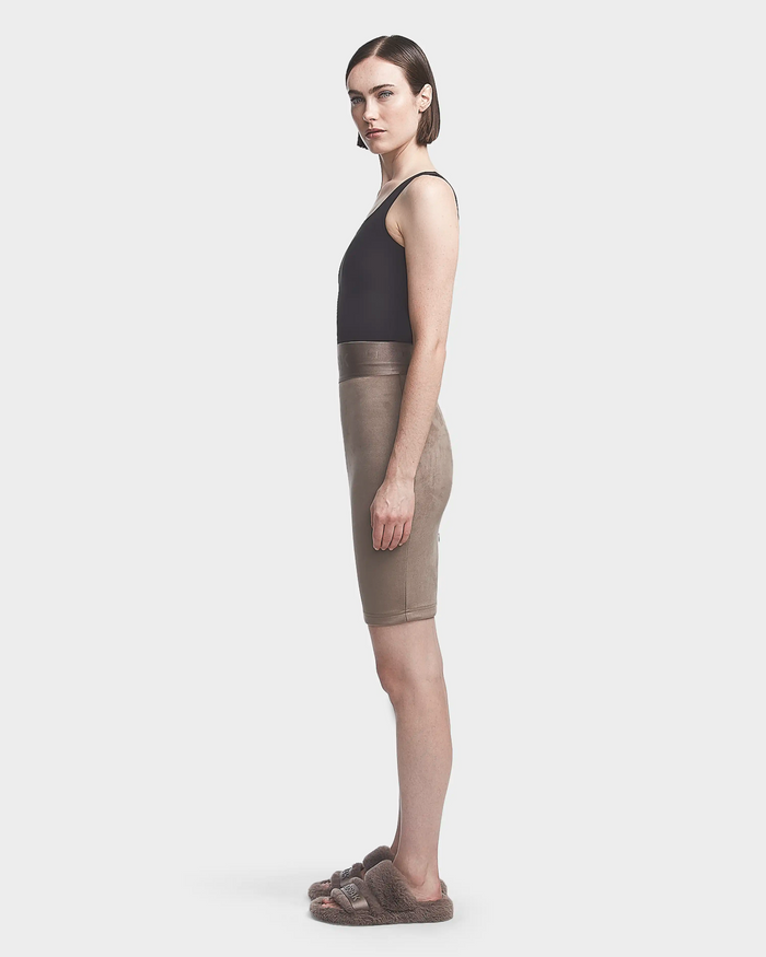 A woman in profile wears the Rudsak CHEYNA - 8523514 Cement faux suede pencil skirt, a black sleeveless top, and fuzzy brown slide sandals, standing against a plain light gray background.