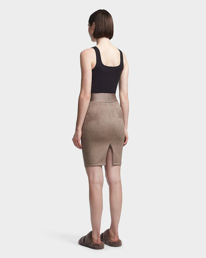 A person with short brown hair wears a black sleeveless top and the Rudsak CHEYNA - 8523514 Cement womens faux suede pencil skirt with a back slit, paired with brown fuzzy slippers, standing against a plain light background.