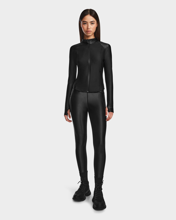 A woman faces forward, wearing Rudsaks CLARE - 8524519 Black fitted zip-up jacket and matching shiny stretch leggings with black boots, against a plain white background that highlights her sculpted silhouette.